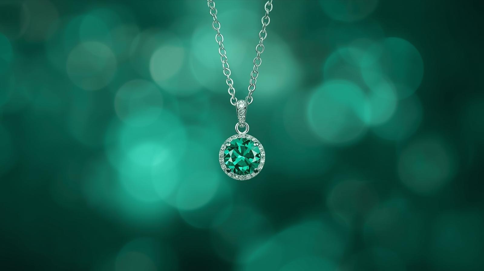 A round emerald pendant haloed with pavé diamonds, suspended on a white-gold chain over a green bokeh ground.